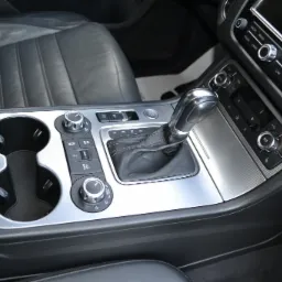 Wnętrze samochodu z widocznym tunelem środkowym, dźwignią zmiany biegów, panelem sterowania klimatyzacją i radiem oraz uchwytami na napoje. Skórzana tapicerka fotela kierowcy widoczna w lewym...