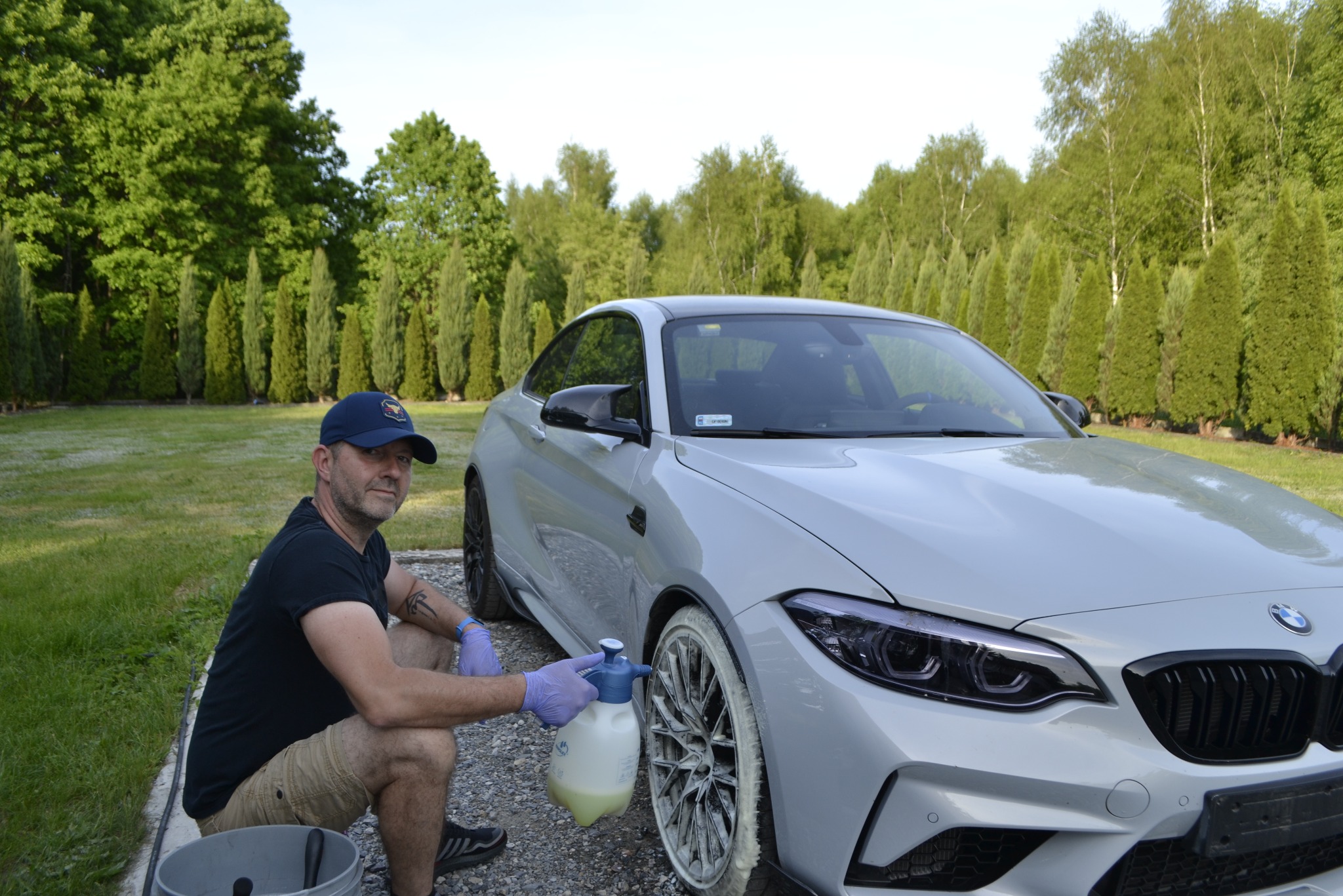 Mężczyzna w niebieskiej czapce i rękawicach ochronnych, klęczy obok srebrnego BMW M2, aplikując pianę z rozpylacza na felgi, na tle zielonego trawnika i rzędu drzew.