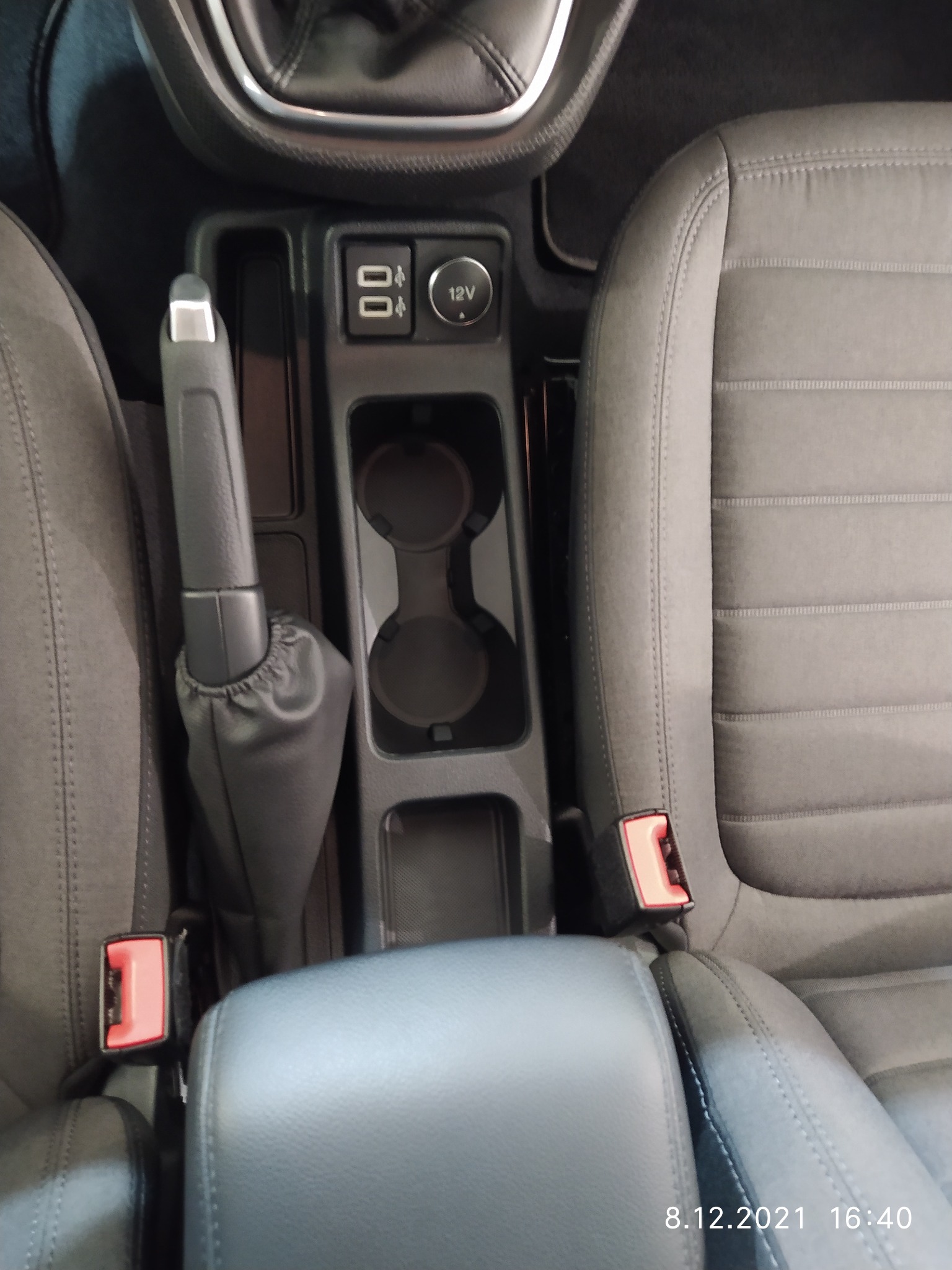 Wnętrze samochodu: konsola środkowa z dźwignią hamulca ręcznego, uchwytami na napoje, portami USB i gniazdem 12V, obok fotel z szarej tapicerki z widocznym zapięciem pasów.