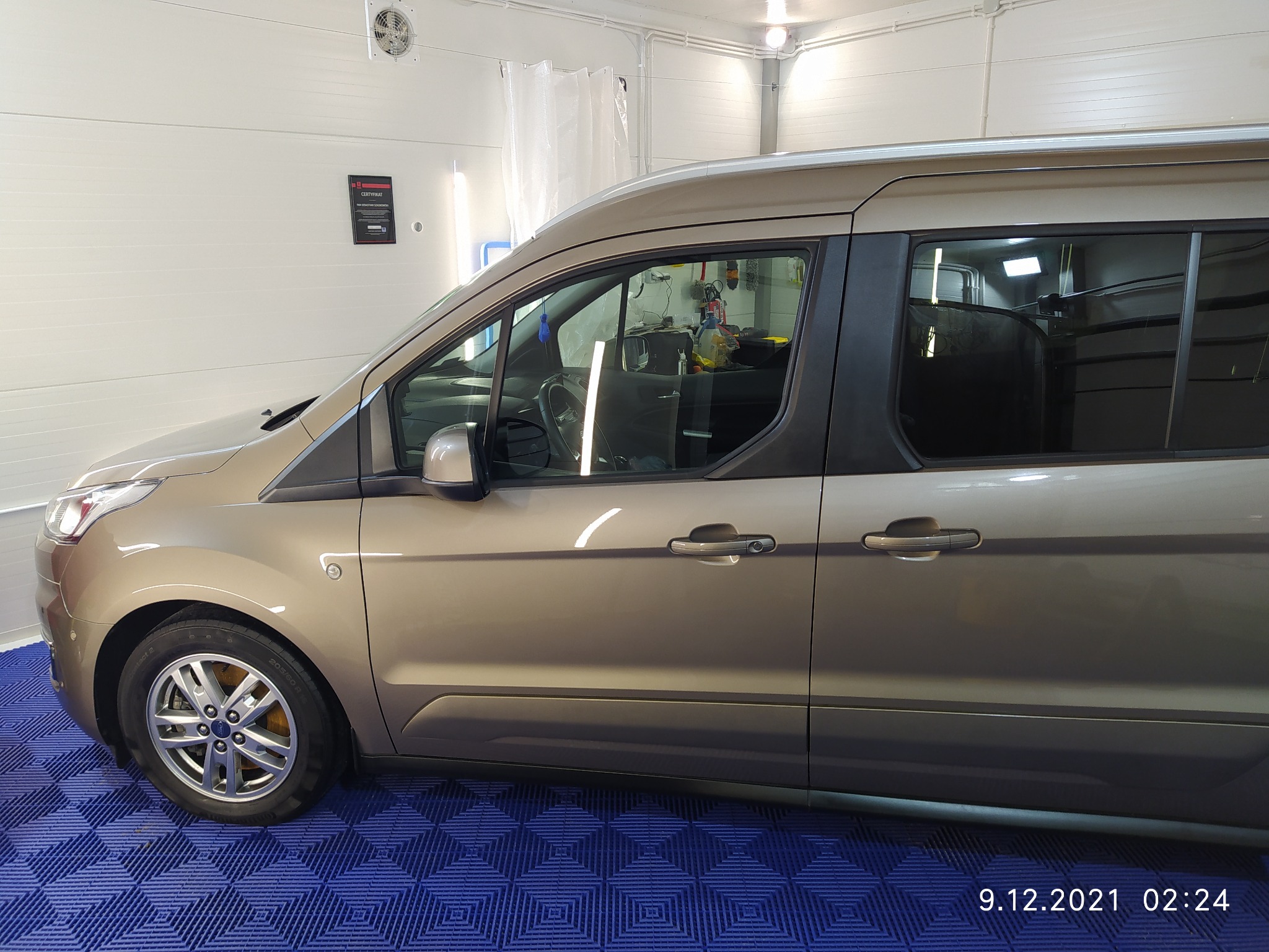 Złoty van Ford Transit w jasnym pomieszczeniu myjni, widoczne niebieskie podłoże z wzorem i certyfikat na ścianie.