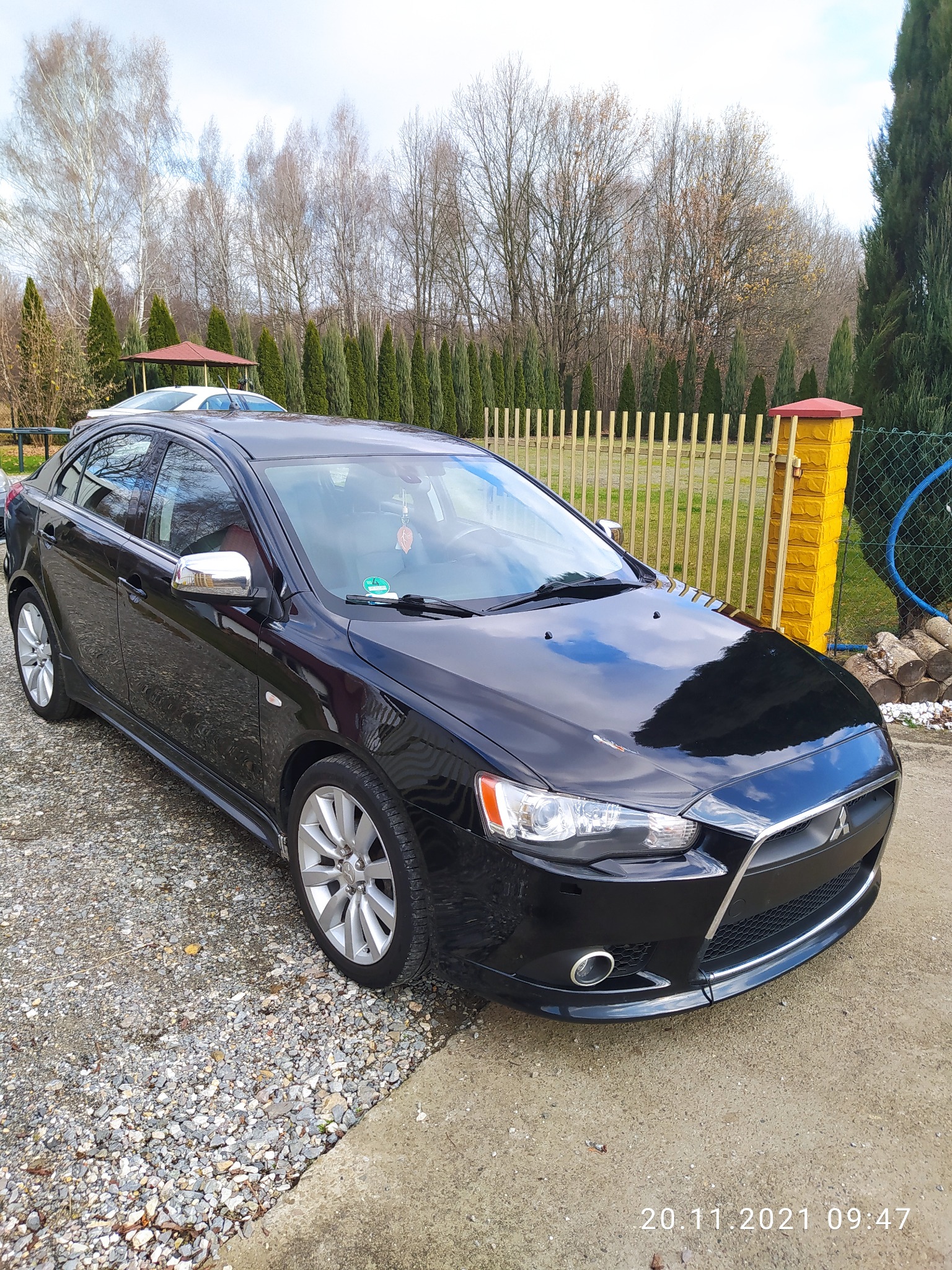 Błyszczący, czarny samochód osobowy marki Mitsubishi Lancer, zaparkowany na żwirowym podjeździe przed domem z drewnianym płotem i zielonymi tujami w tle.