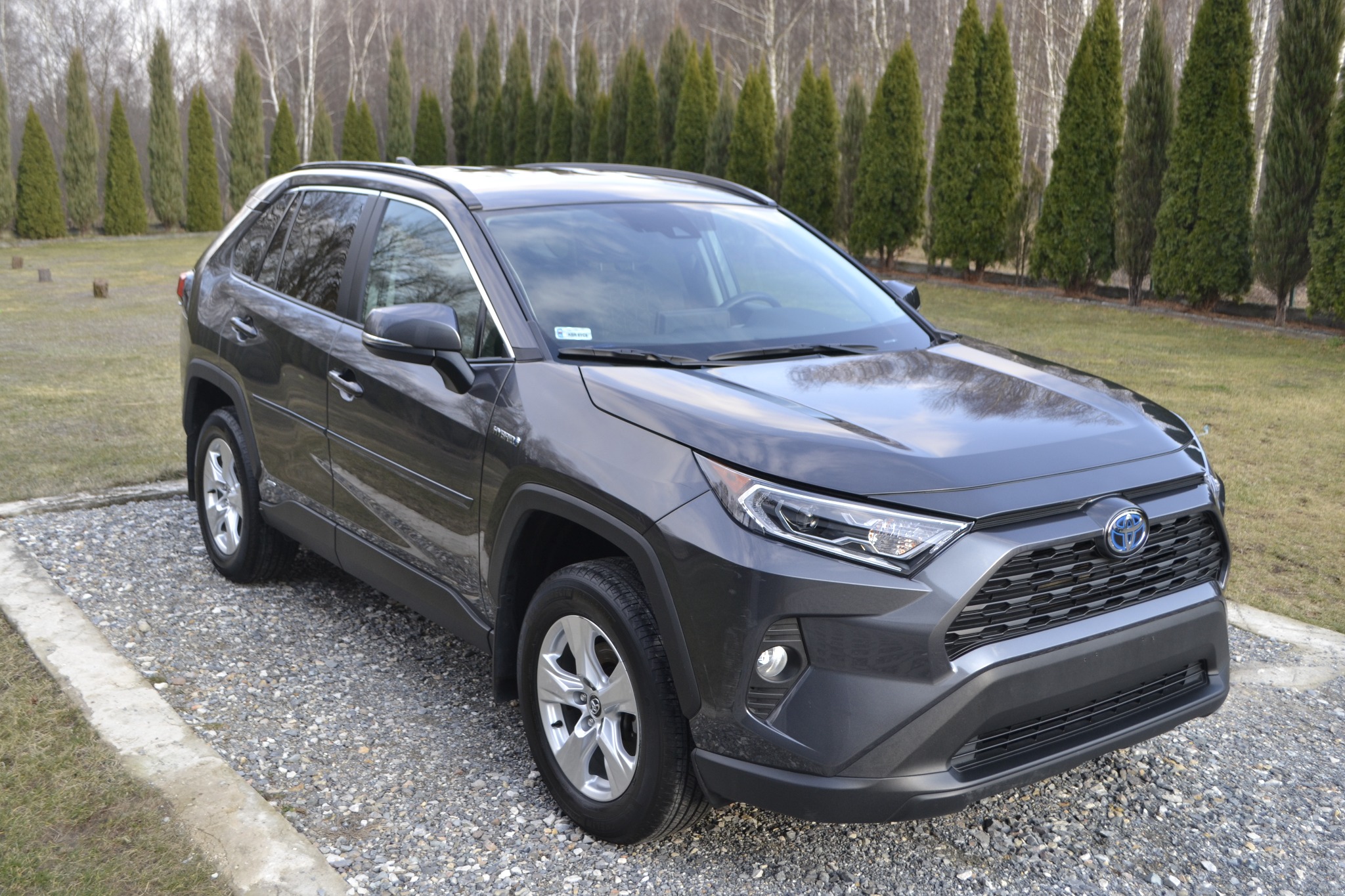 Szary samochód typu SUV, Toyota RAV4 Hybrid, zaparkowany na żwirowym podjeździe przed rzędem wysokich, zielonych drzew.