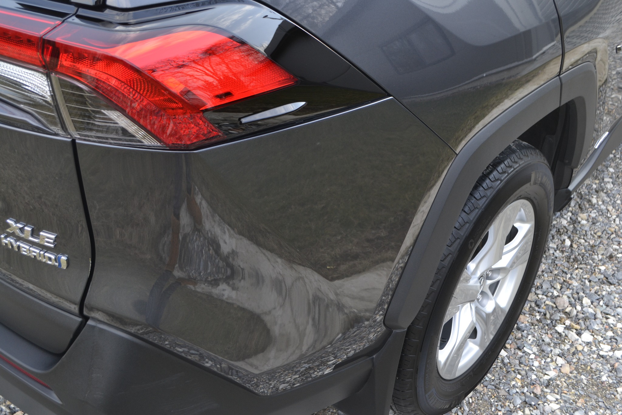 Fragment tylnej części szarego samochodu XLE Hybrid, widoczny błotnik, opona i światło z odbiciem drzew, stojącego na żwirowym podłożu.