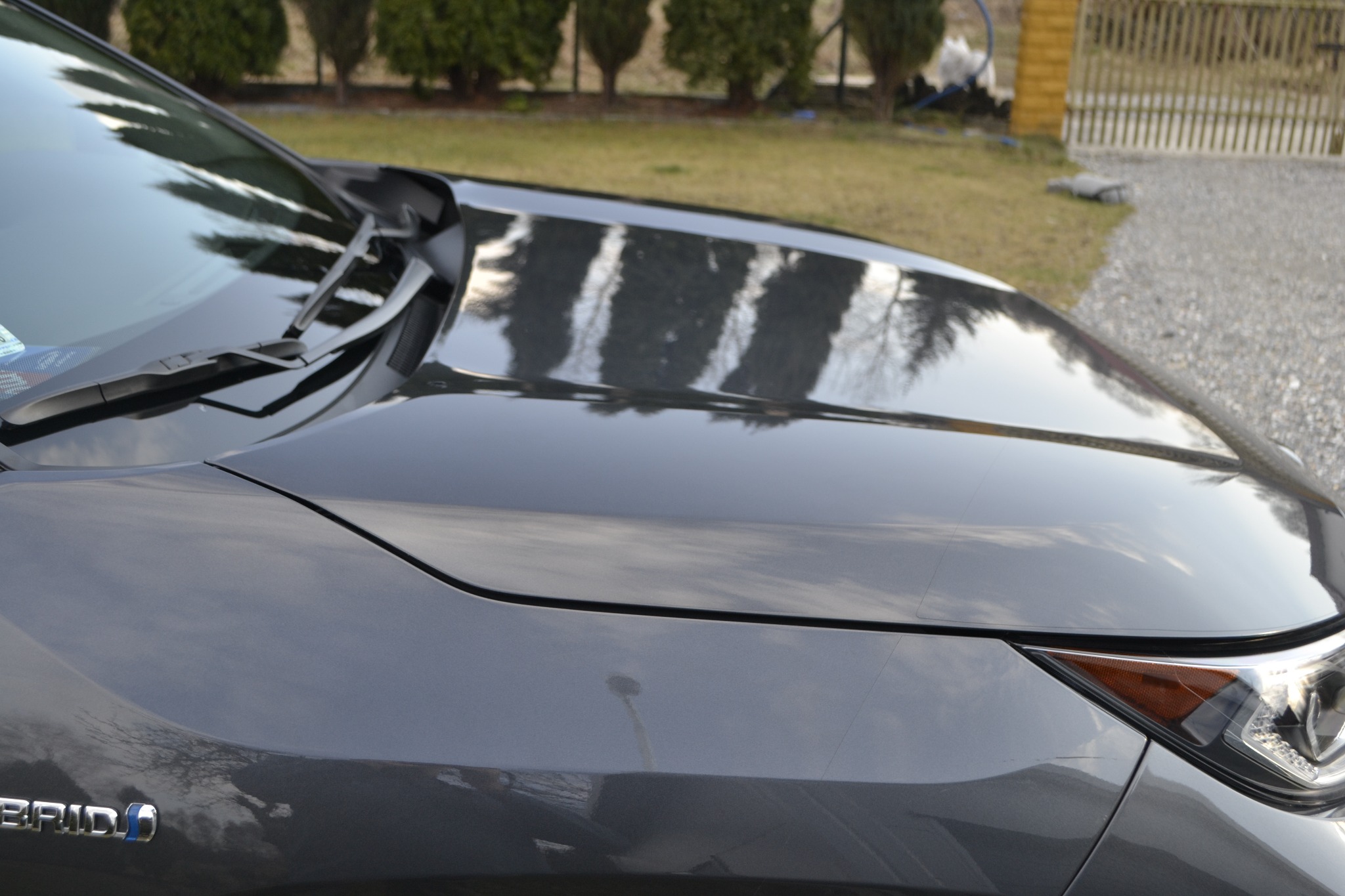 Błyszcząca maska szarego samochodu hybrydowego z widocznymi refleksami drzew i nieba, zarys przedniego reflektora.