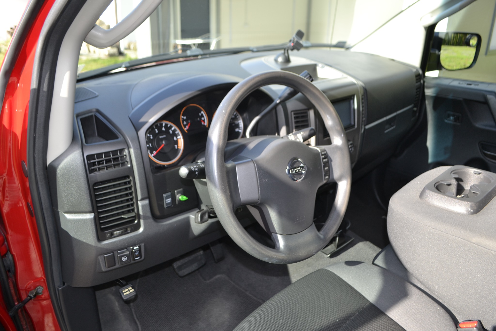 Wnętrze czerwonego samochodu Nissan Titan z widoczną deską rozdzielczą, kierownicą z logo Nissan i szarą tapicerką foteli. Widoczne detale: kratka wentylacyjna, wskaźniki na desce rozdzielczej...