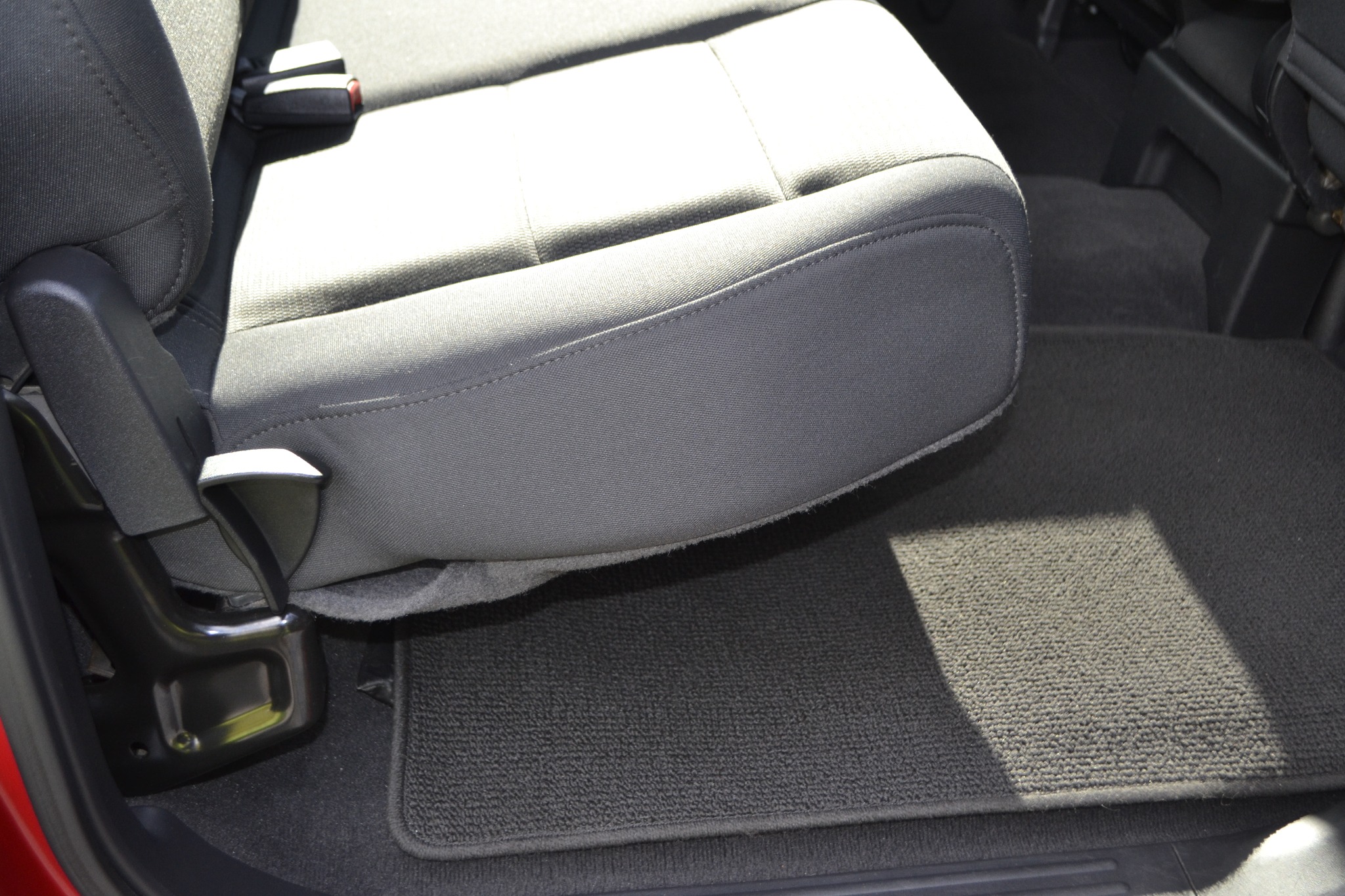 Fragment wnętrza samochodu: szary fotel z zagłówkiem i pasami bezpieczeństwa oraz dywanik podłogowy, oświetlone jasnym światłem słonecznym.