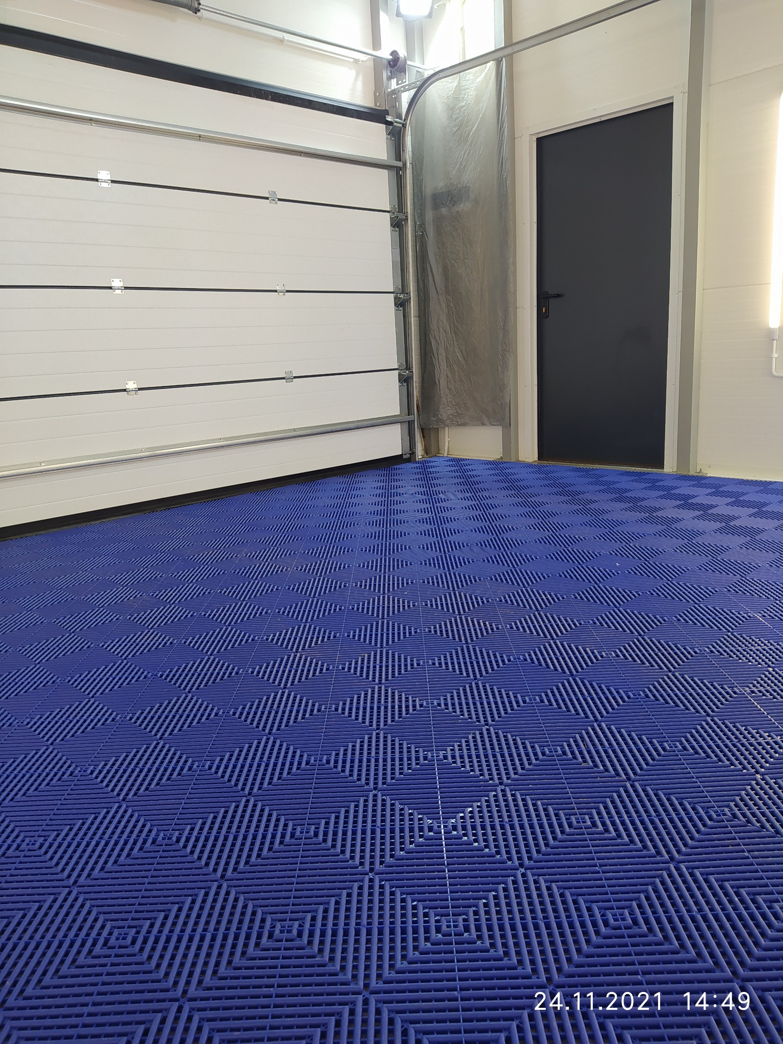 Wnętrze myjni samochodowej z niebieską podłogą z tworzywa sztucznego i otwartą białą bramą garażową.