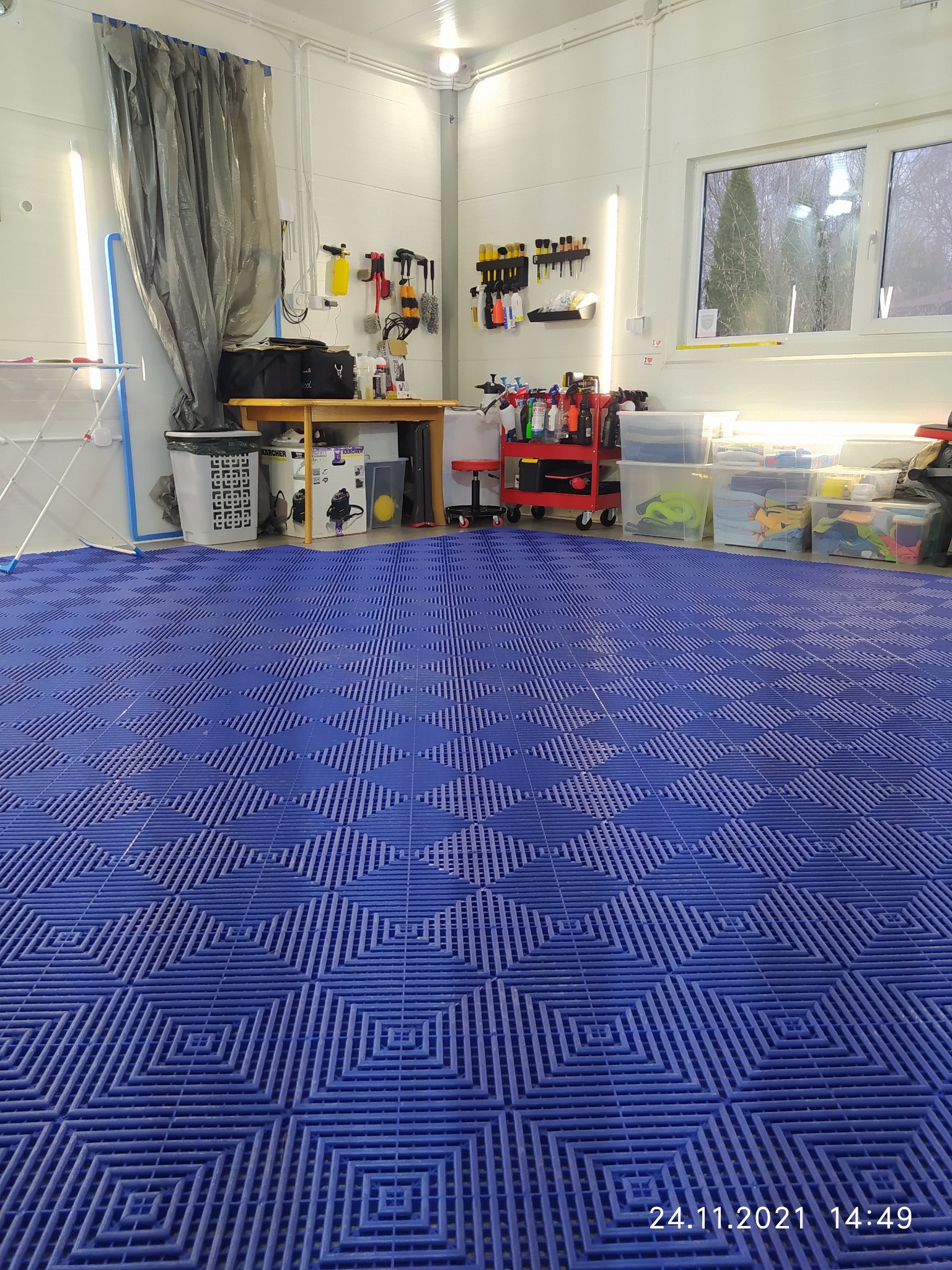 Wnętrze myjni samochodowej z niebieską podłogą z tworzywa sztucznego, narzędziami i akcesoriami do czyszczenia aut na półkach i w wózkach, widok z perspektywy podłogi.