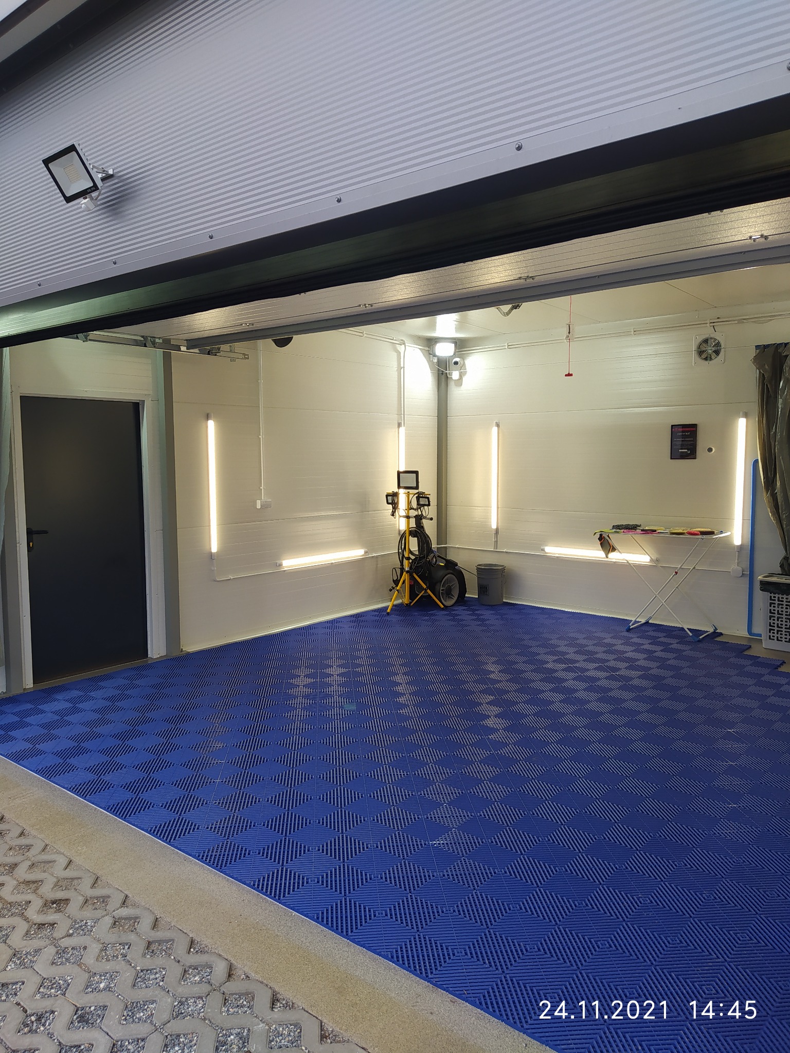 Wnętrze myjni samochodowej z niebieską podłogą, oświetleniem LED i sprzętem do czyszczenia, widoczne z zewnątrz przez otwarte drzwi garażowe.