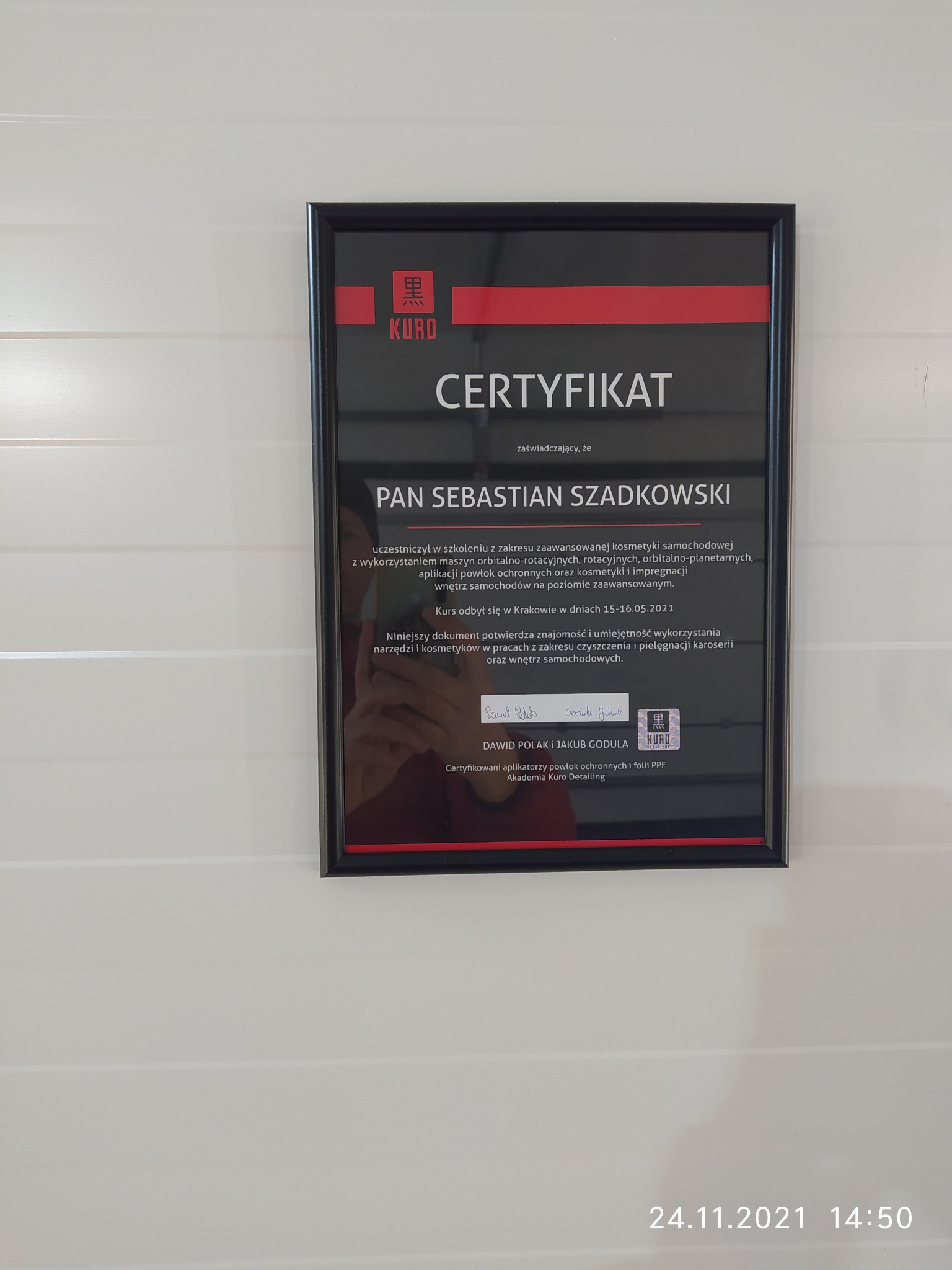Certyfikat Akademii Kuro Detailing dla Pana Sebastiana Szadkowskiego potwierdzający ukończenie szkolenia z zaawansowanej kosmetyki samochodowej, w czarnej ramce na białej ścianie.