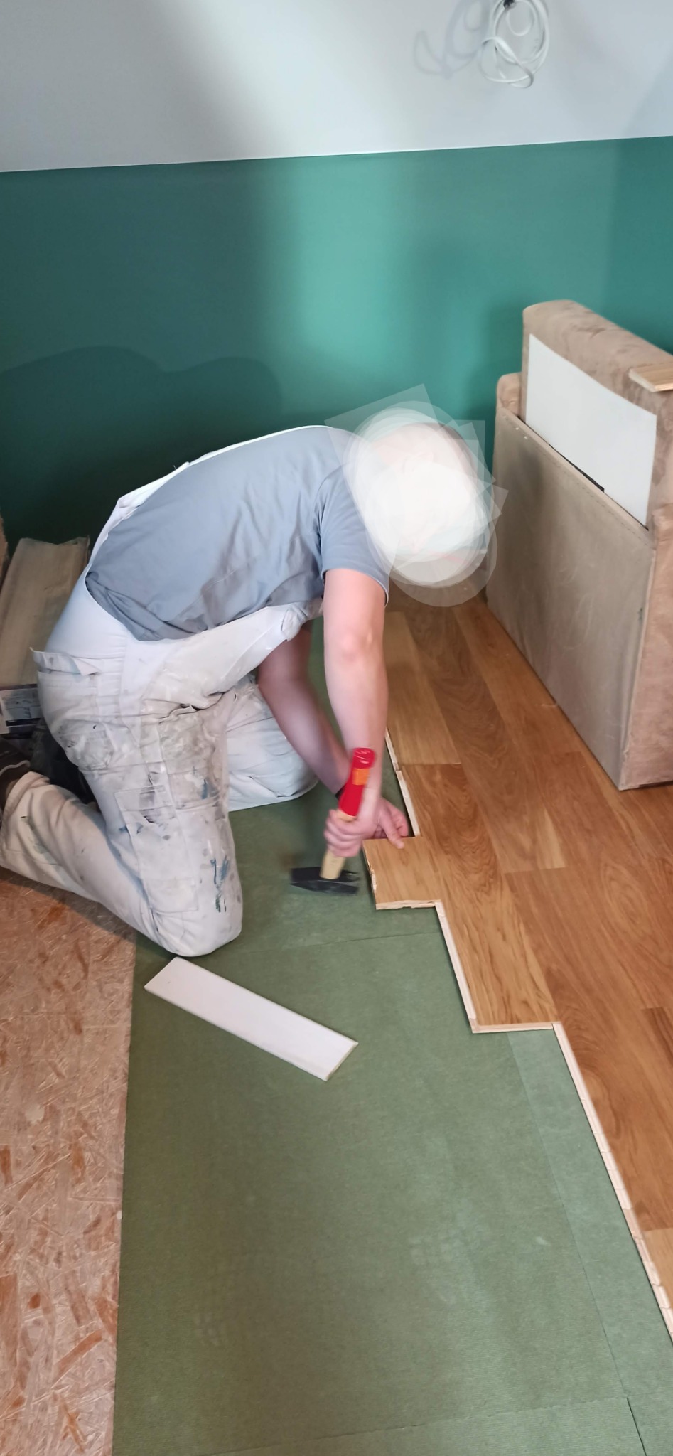 Pracownik w ubraniu roboczym dobija młotkiem panel podłogowy w rogu pomieszczenia, widoczna zielona podkładka pod panele i fragment listwy przypodłogowej.