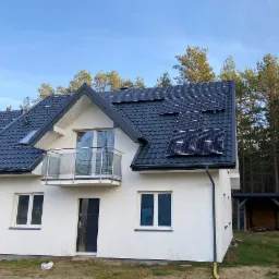 Nowoczesny dom jednorodzinny z szarą dachówką i panelami fotowoltaicznymi na dachu, widok z zewnątrz w słoneczny dzień, na tle lasu.