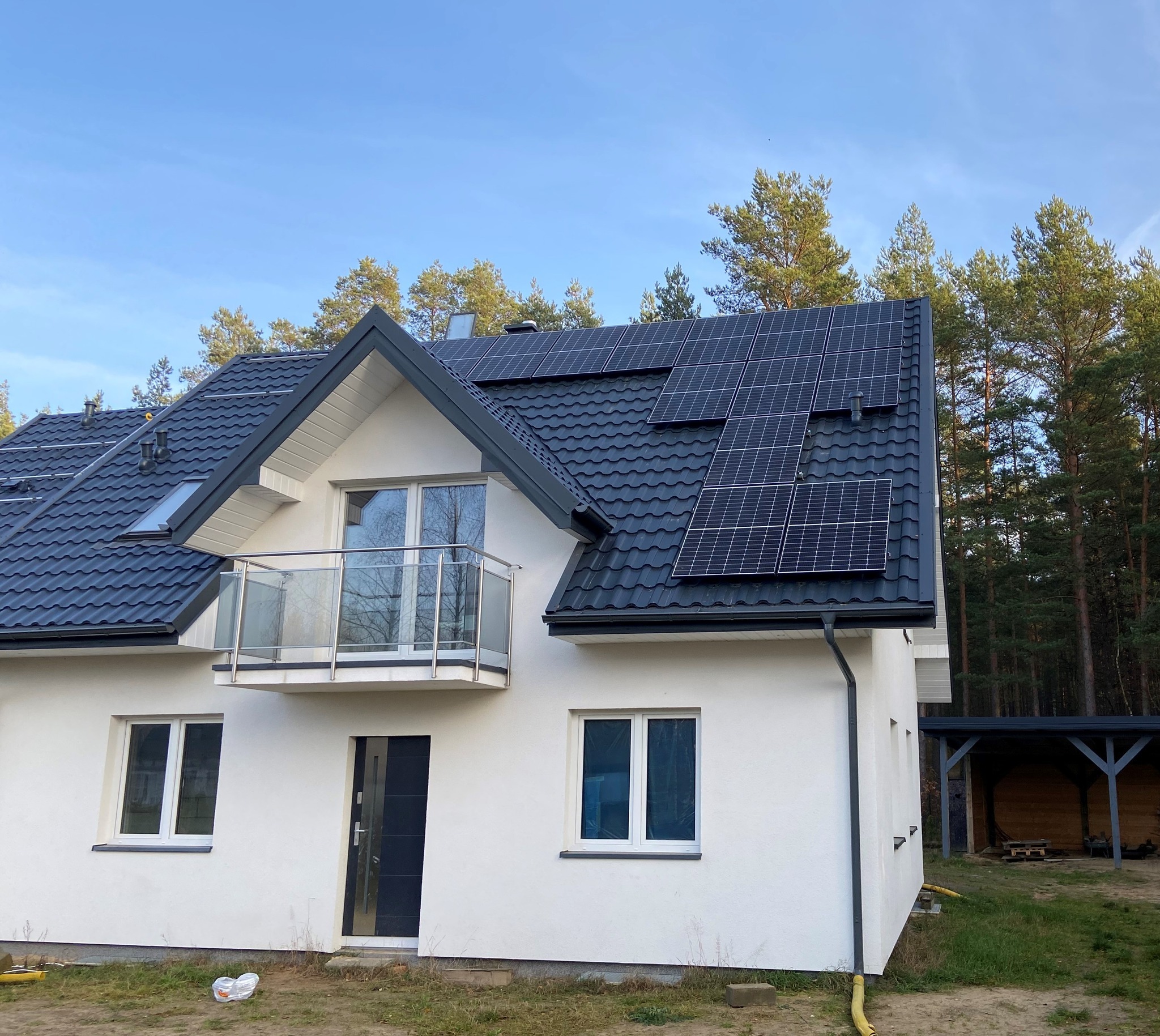 Nowoczesny dom jednorodzinny z szarą dachówką i panelami fotowoltaicznymi na dachu, widok z zewnątrz w słoneczny dzień, na tle lasu.