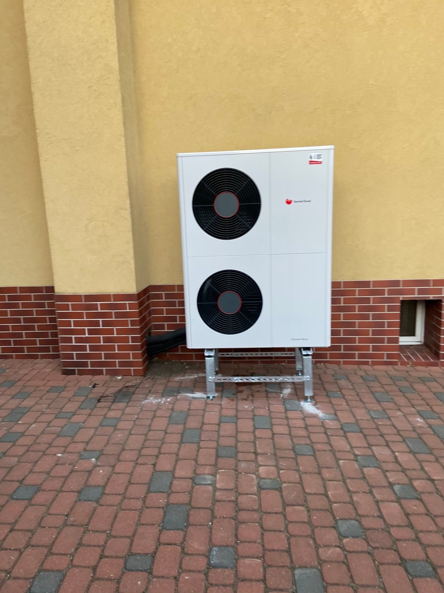 Biała jednostka zewnętrzna z dwoma wentylatorami na metalowej konstrukcji, zamontowana przy elewacji budynku z ceglanym cokołem i żółtym tynkiem.