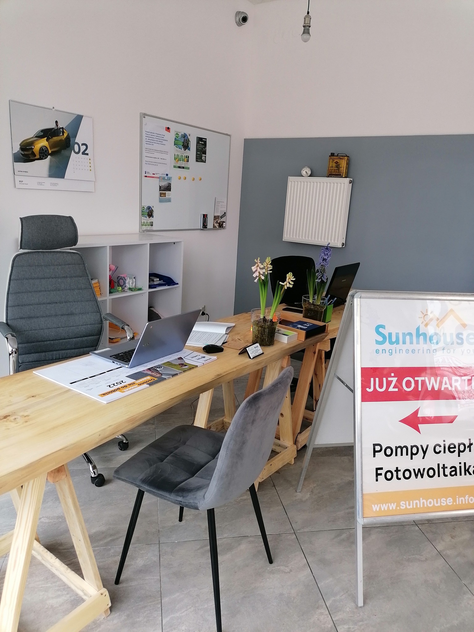 Wnętrze biura firmy Sunhouse z drewnianym biurkiem, laptopami, kwiatami i tablicą reklamową informującą o otwarciu i ofercie na pompy ciepła i fotowoltaikę.