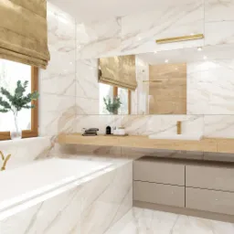 Jasna łazienka z marmurowymi płytkami, wanna przy oknie z roletą rzymską, podwójna umywalka na drewnianym blacie i złota armatura.