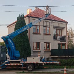 Podnośnik koszowy Liftmar Marek Rybowski - Podnośnik koszowy Euro B 20 T na pojeździe ustawiony przed dwupiętrowym domem z czerwonym dachem, z osobą w koszu na wysokości dachu, prawdopodobnie podczas prac konserwacyjnych.