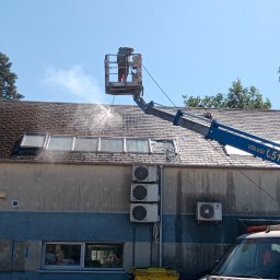 Podnośnik koszowy Liftmar Marek Rybowski - Pracownik w podnośniku koszowym czyści dach budynku z mchu i zabrudzeń za pomocą myjki ciśnieniowej; widoczne okna dachowe, komin i fragment elewacji.