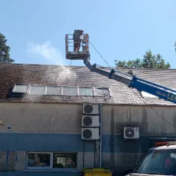 Pracownik w podnośniku koszowym czyści dach budynku z mchu i zabrudzeń za pomocą myjki ciśnieniowej; widoczne okna dachowe, komin i fragment elewacji.