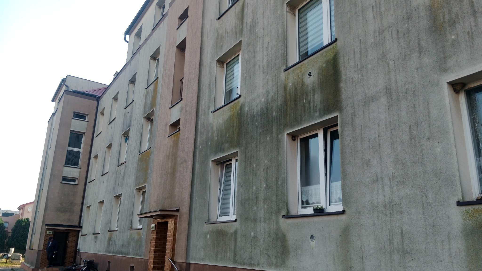 Szary budynek wielorodzinny z widocznymi zabrudzeniami i zaciekami na elewacji, z białymi oknami i numerami na wejściach.