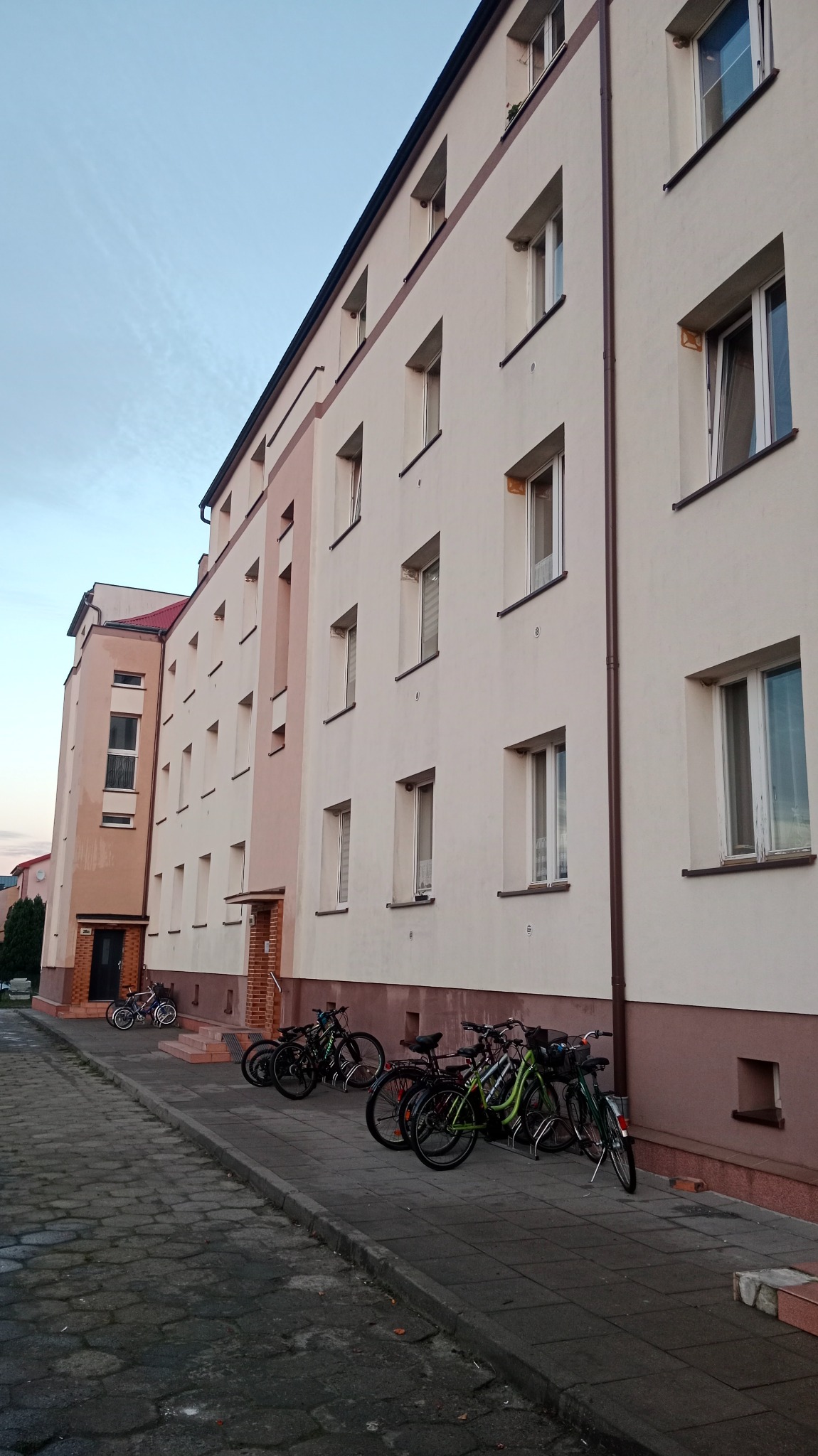 Wielopiętrowy budynek mieszkalny z beżową elewacją, widoczne okna z brązowymi ramami, rynny i zaparkowane rowery na chodniku z kostki brukowej.