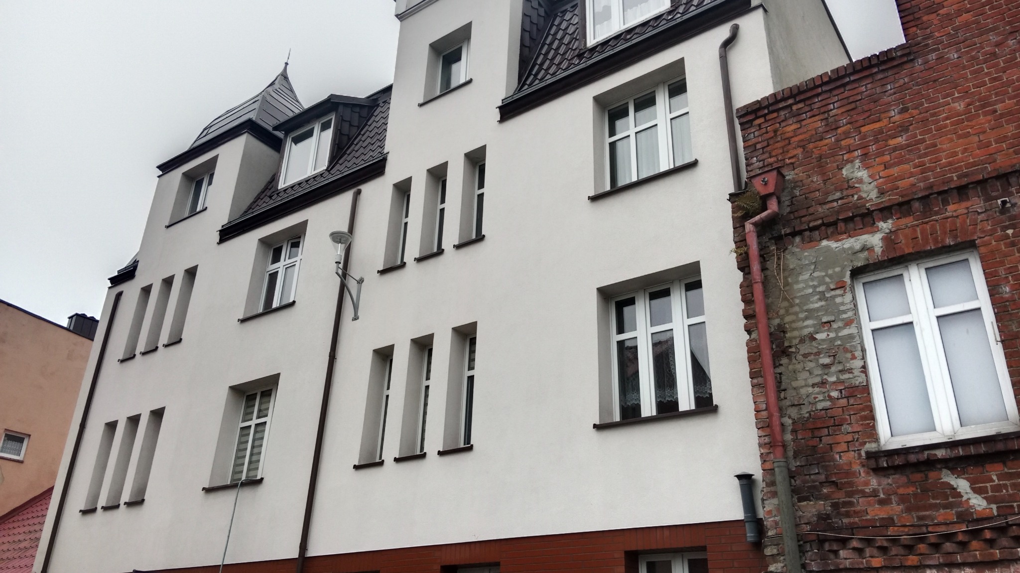 Wielopiętrowy budynek z białą elewacją i ciemnym dachem, widoczny z dołu, sąsiadujący z budynkiem z czerwonej cegły z rynną. Na elewacji widoczne pionowe wgłębienia i lampa oświetleniowa.