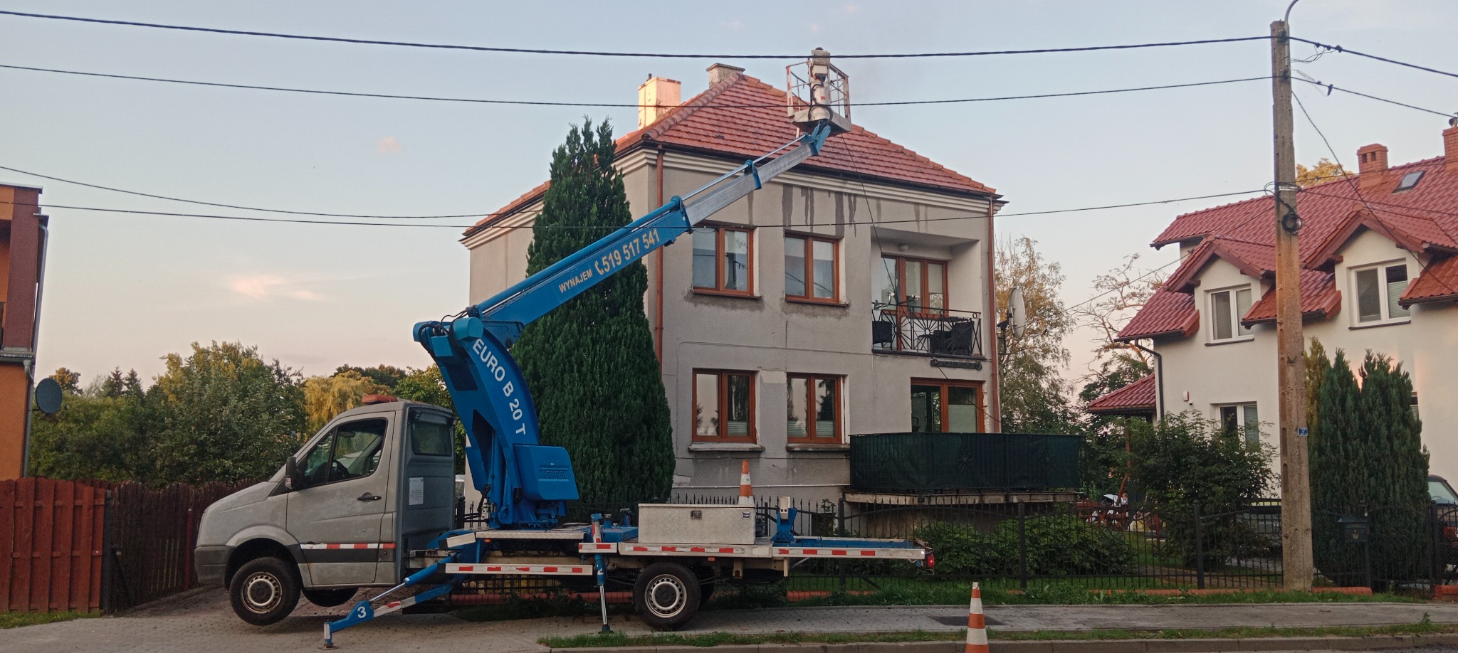 Podnośnik koszowy Euro B 20 T na pojeździe ustawiony przed dwupiętrowym domem z czerwonym dachem, z osobą w koszu na wysokości dachu, prawdopodobnie podczas prac konserwacyjnych.