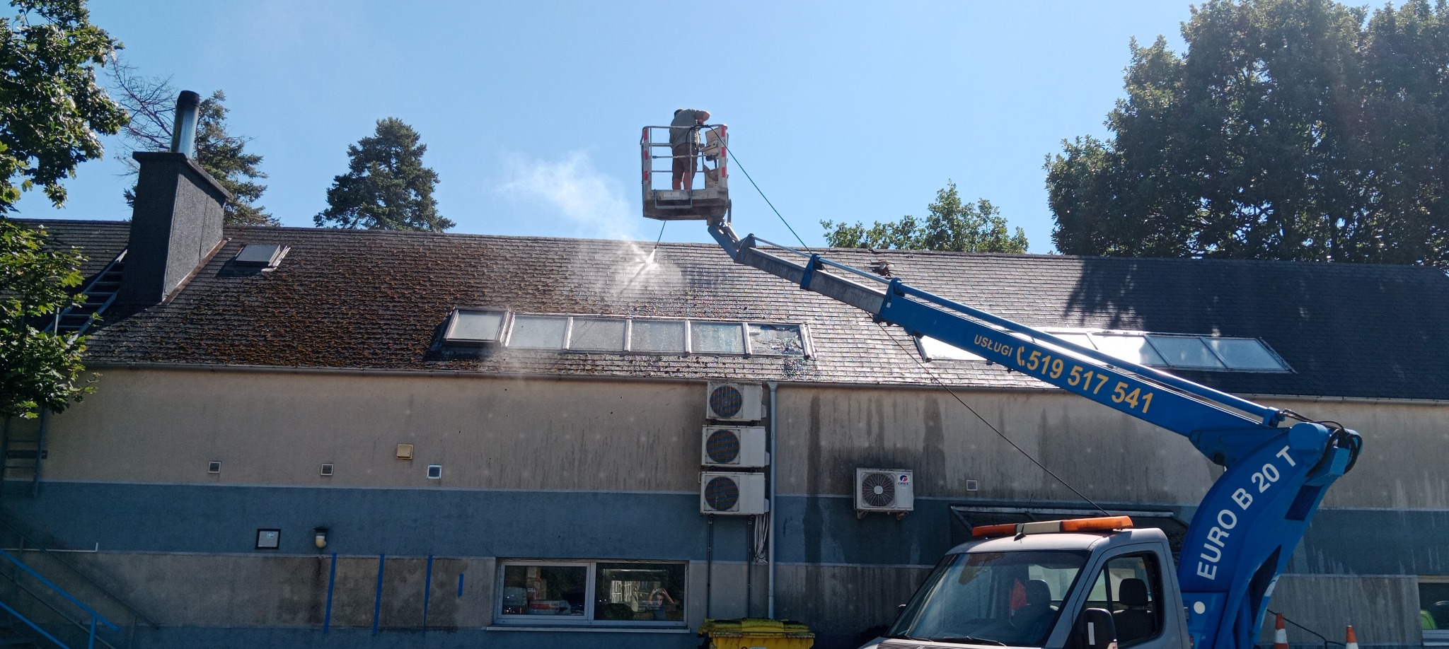 Pracownik w podnośniku koszowym czyści dach budynku z mchu i zabrudzeń za pomocą myjki ciśnieniowej; widoczne okna dachowe, komin i fragment elewacji.