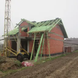 Budowa domu z czerwonej cegły: ściany wzniesione, zielona więźba dachowa, dźwig budowlany obok. Widok na plac budowy w pochmurny dzień.