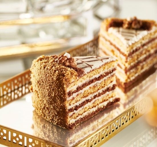 Dwa apetyczne kawałki wielowarstwowego tortu miodowego na złotej, ażurowej tacy, z orzechami włoskimi na wierzchu i delikatnym kremem, oświetlone ciepłym światłem.