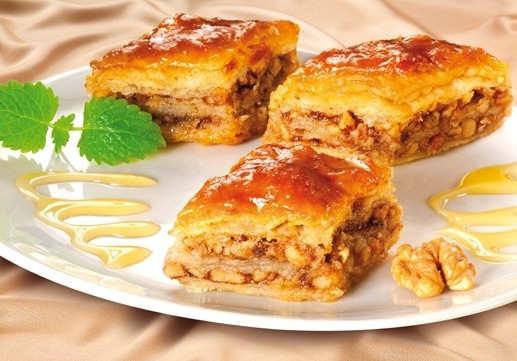 Trzy kawałki baklawy z orzechami na białym talerzu, polane syropem i ozdobione listkiem mięty oraz połówką orzecha włoskiego, na beżowym tle.