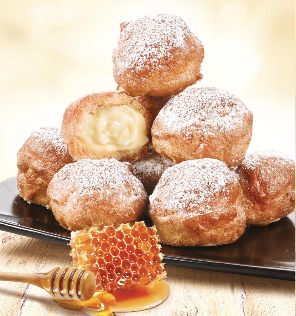 Piramide z pączków posypanych cukrem pudrem, jeden z kremem waniliowym, obok plaster miodu i nabierak z kapiącym miodem na czarnym talerzu.