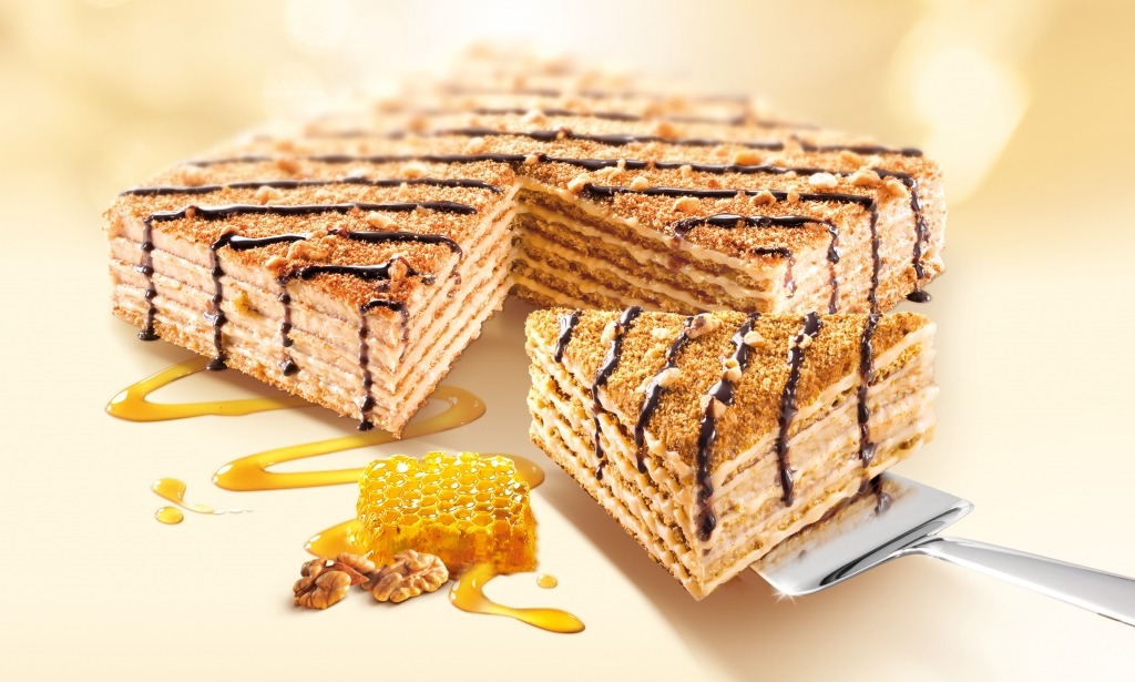 Wielowarstwowy tort miodowy z orzechami, polany czekoladą, plaster miodu i orzechy włoskie obok, podany na metalowej łopatce.