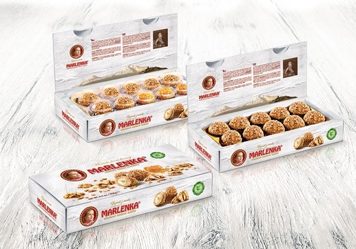 Trzy pudełka ciastek Marlenka na białym, drewnianym tle. Dwa pudełka są otwarte, prezentując kuliste ciastka o orzechowej posypce. Na opakowaniach widoczne logo z portretem i napisem Marlenka.