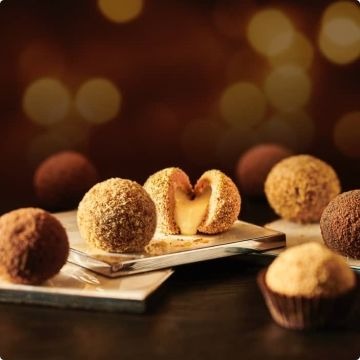 Elegancko zaaranżowane praliny truflowe o różnych smakach i posypkach, jedna przekrojona ukazująca kremowe wnętrze, ułożone na metalowej tacy na ciemnym tle z rozmytymi, ciepłymi światłami.