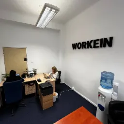 Wnętrze biura z dwoma pracownikami przy biurkach, logo 'WORKEIN' na ścianie, dystrybutor wody i ciemna wykładzina podłogowa.