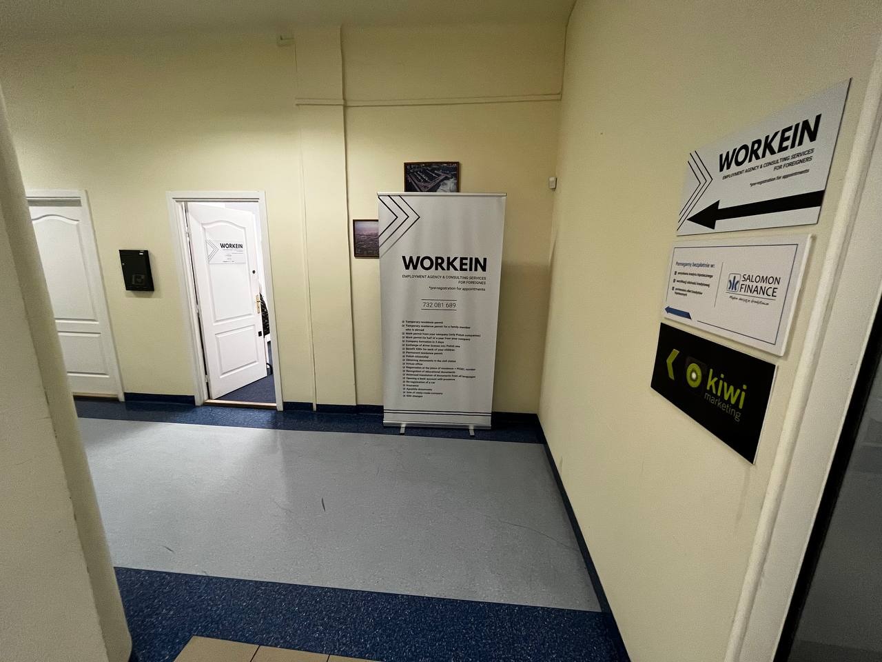 Wnętrze biura z plakatem i tabliczkami informacyjnymi firm Workein, Salomon Finance i Kiwi Marketing na ścianie, widoczne drzwi.