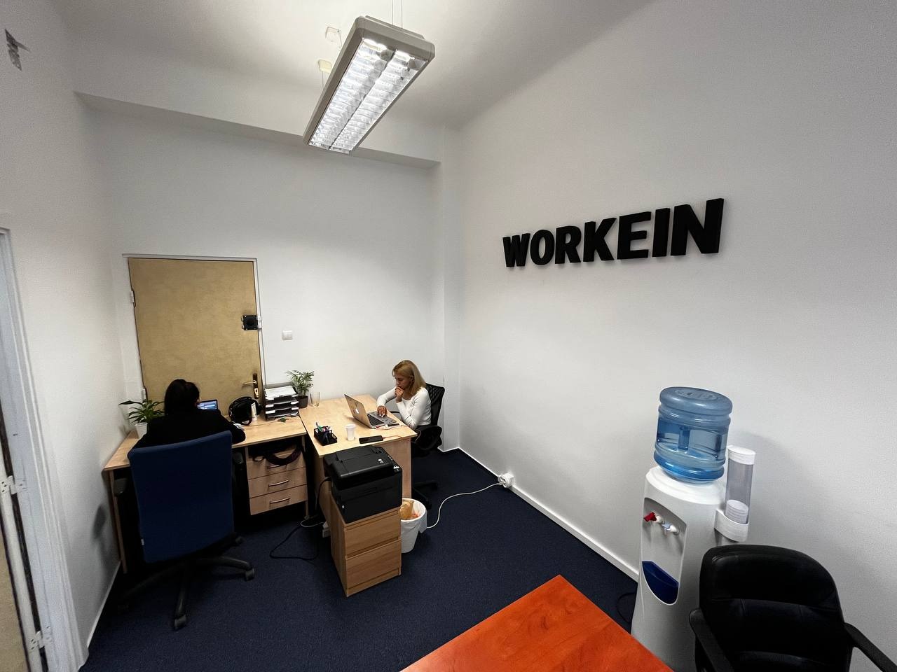 Wnętrze biura z dwoma pracownikami przy biurkach, logo 'WORKEIN' na ścianie, dystrybutor wody i ciemna wykładzina podłogowa.