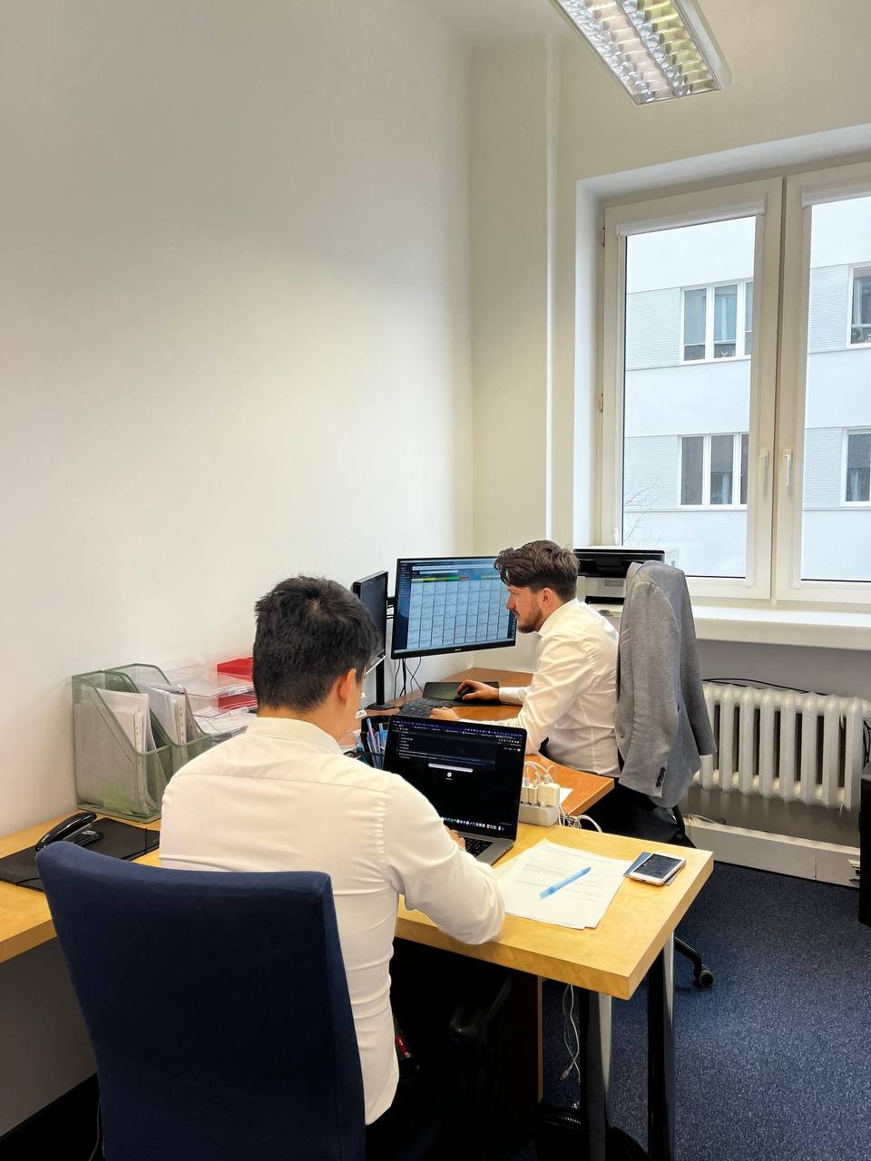 Dwóch mężczyzn w białych koszulach pracuje przy biurkach w jasnym biurze, jeden z laptopem, drugi z dwoma monitorami wyświetlającymi kalendarz, szara marynarka zawieszona na krześle.