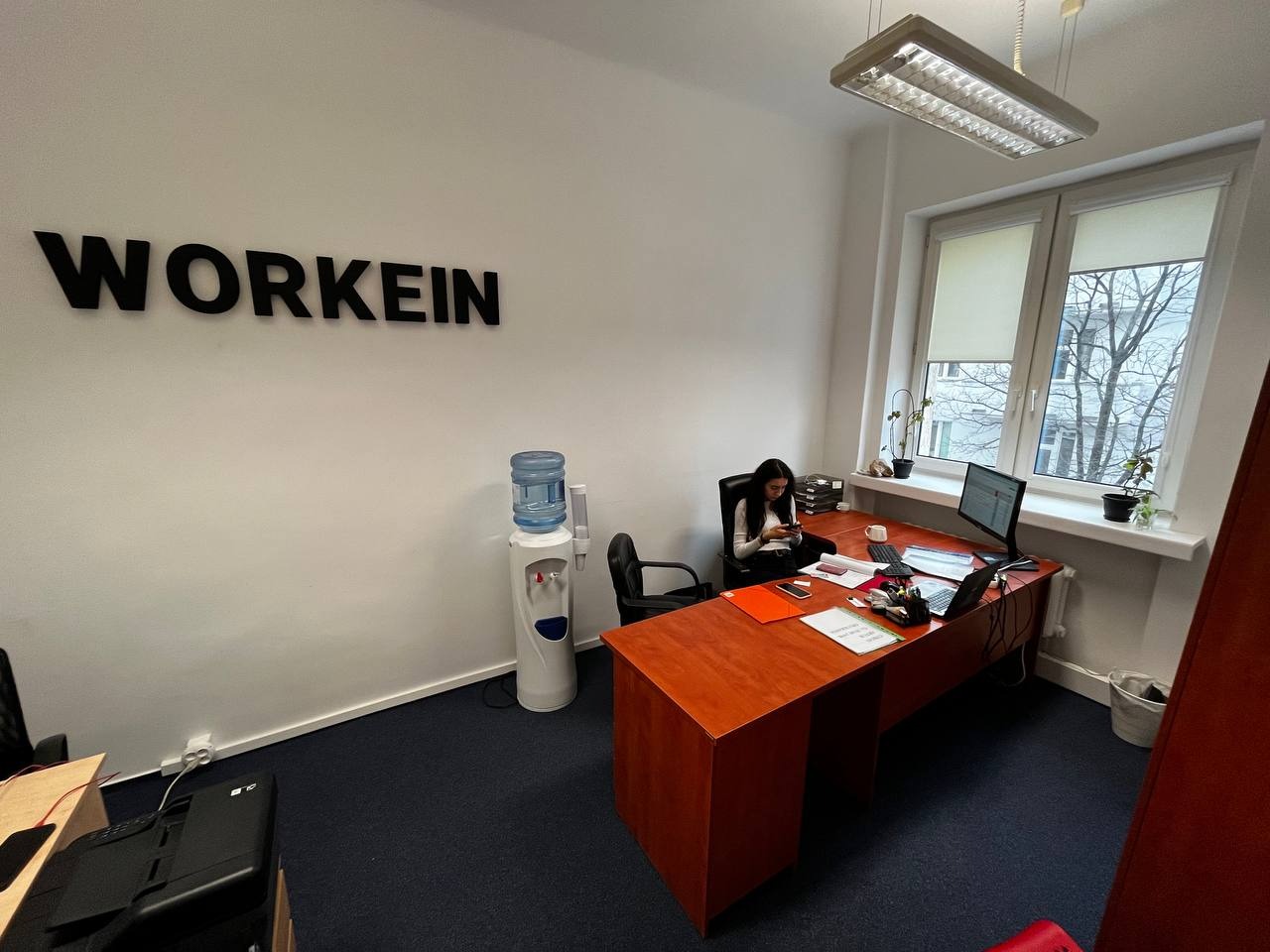 Wnętrze biura z biurkiem, komputerem i siedzącą kobietą, na ścianie napis WORKEIN, dystrybutor wody i okno z widokiem na drzewa.