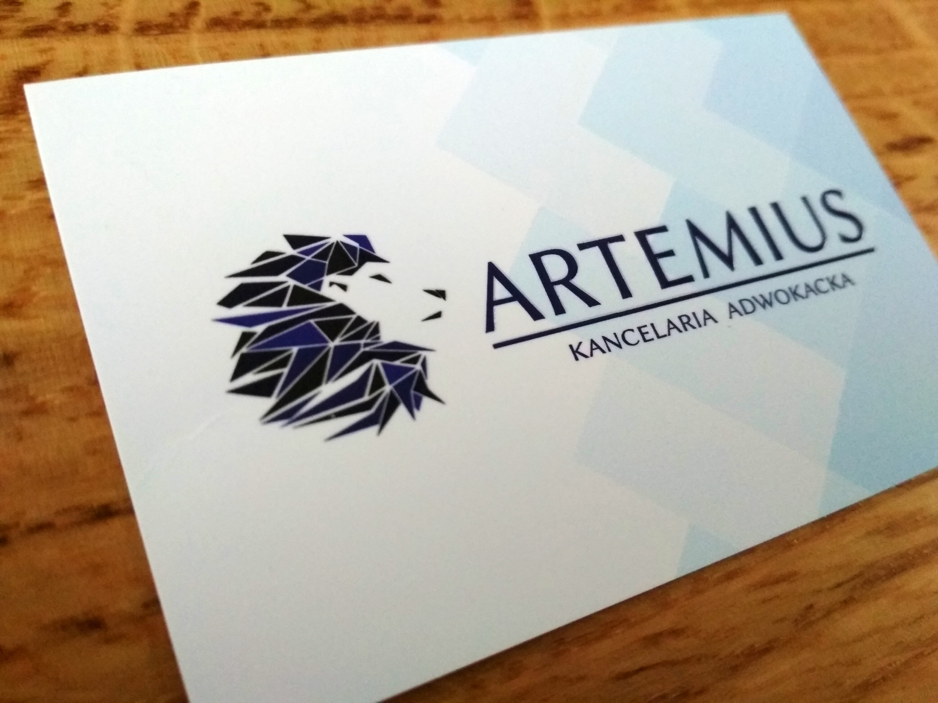Wizytówka z logo kancelarii w formie geometrycznej głowy lwa w kolorach granatu i czerni, nazwa firmy 'ARTEMIUS KANCELARIA ADWOKACKA' zapisana ciemnogranatową czcionką.