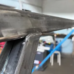 Szczegółowe ujęcie spoiny łączącej dwa metalowe profile, widoczne ślady obróbki i minimalne niedoskonałości spoiny, w tle warsztat.