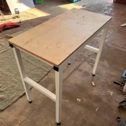 Stół warsztatowy z metalową ramą i drewnianym blatem, widoczne ślady użytkowania na podłodze warsztatu, oświetlenie dzienne.