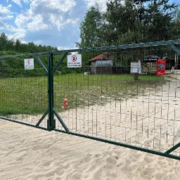 Zielona brama z siatki na teren strzelnicy, z tabliczkami ostrzegawczymi o zakazie wstępu, piasek na ziemi, w tle budynek i drzewa.