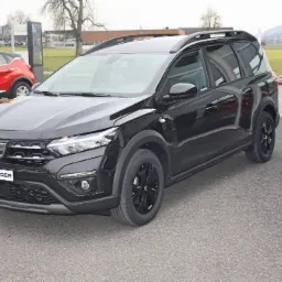 Czarny samochód typu kombi Dacia Jogger z relingami dachowymi, zaparkowany na zewnątrz salonu samochodowego, z widocznym czerwonym autem w tle.