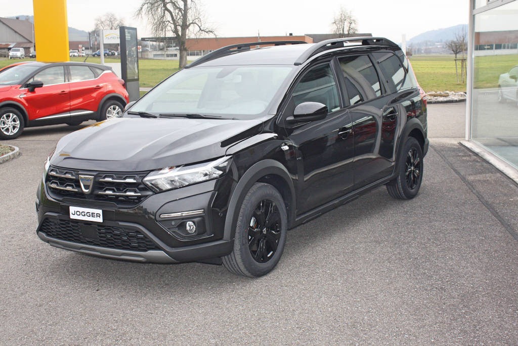 Czarny samochód typu kombi Dacia Jogger z relingami dachowymi, zaparkowany na zewnątrz salonu samochodowego, z widocznym czerwonym autem w tle.