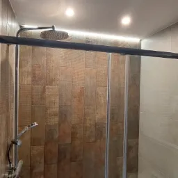 Kabina prysznicowa z deszczownicą i brązowymi płytkami imitującymi drewno, oświetlenie LED nad kabiną, widoczny odpływ liniowy w podłodze.