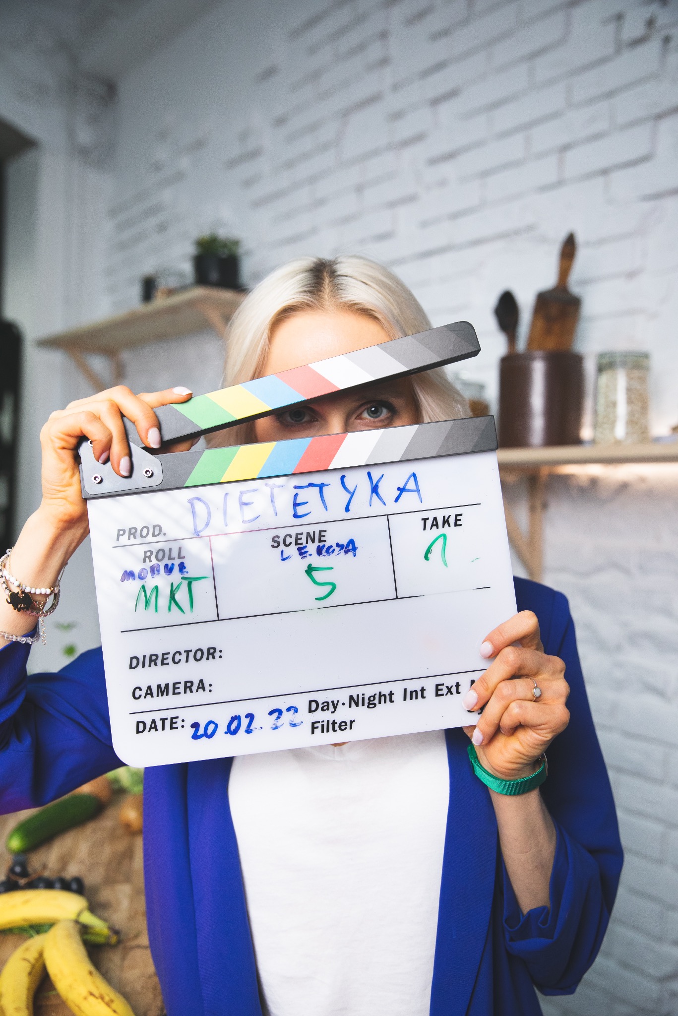 Blondynka z niebieskimi oczami trzyma klaps filmowy z napisem 'Dietetyka', widoczne owoce i warzywa, biała ceglana ściana w tle.