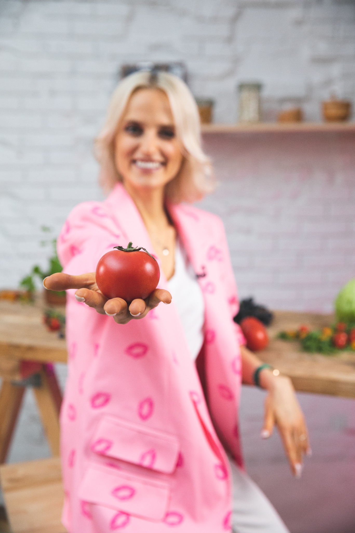 Uśmiechnięta blondynka w różowym żakiecie w usta prezentuje czerwonego pomidora na otwartej dłoni, na drewnianym stole w tle inne warzywa i biała ceglana ściana.