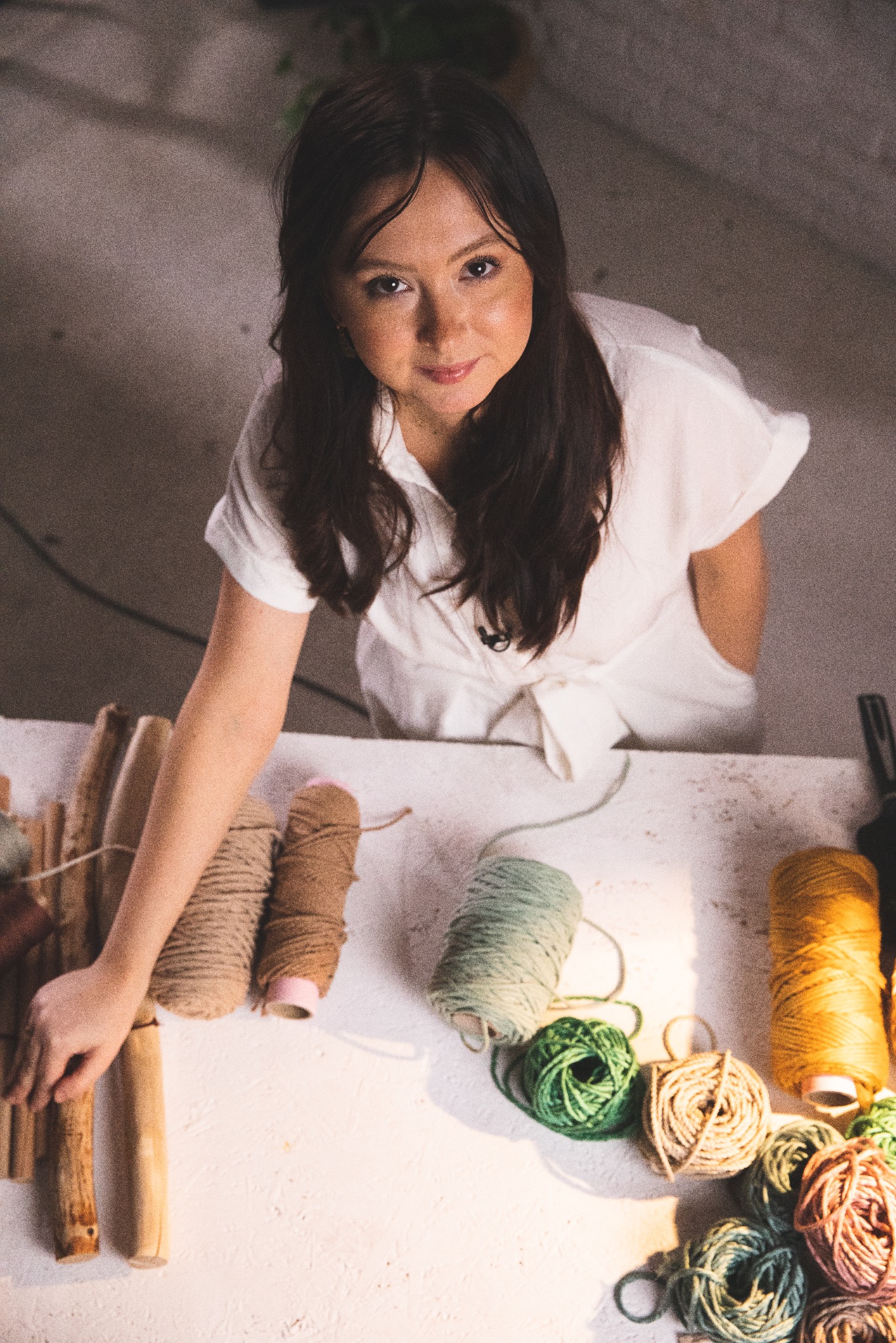 Kobieta w białej sukience, patrząca w górę, pracuje z różnokolorowymi sznurkami i drewnianymi elementami na białym stole, tworząc rękodzieło.